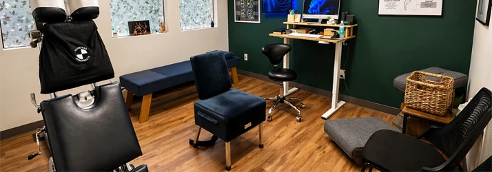 Chiropractic office interior featuring an adjustment table, seating area, workstation, and framed wall displays.